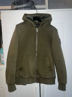 Olivgrön hoodie från Moose Knuckles - Snygg olivgrön hoodie från Moose Knuckles med dragkedja, huva och två stora fickor framtill. Har ett stilrent logomärke på ärmen och ribbade muddar. Perfekt för en avslappnad streetwear-look.