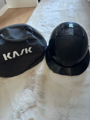 Svart ridhjälm från KASK - Säljer en svart ridhjälm från KASK med blank finish och ventilerande meshdetaljer på ovansidan. Hjälmen har klassisk skärm och kommer med en praktisk svart väska med KASK-logga. Perfekt för dig som vill ha både stil och funktion i stallet.