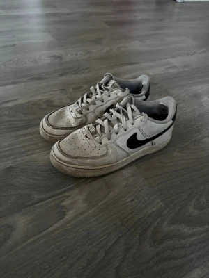 Nike Air Force 1 vita sneakers - Klassiska vita Nike Air Force 1 sneakers med svart swoosh på sidan. Skorna har lågt skaft, perforerad tåbox och är tillverkade i läder med gummisula. Perfekta för dig som gillar streetwear och tidlös design.