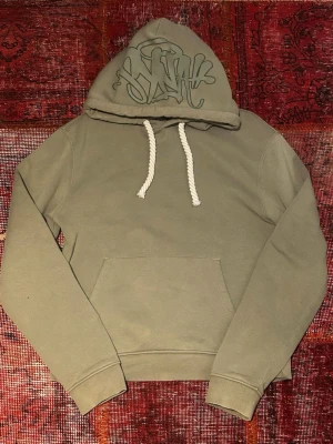 Syna World tracksuit - Säljer en olivgrön hoodie med stor magficka och vita, tjocka dragsnören. Huvan har ett coolt graffiti-inspirerat tryck i samma färgton. Hoodien är i mjukt material och har ribbade muddar vid ärmar och nederkant. Perfekt för en avslappnad streetwear-look.