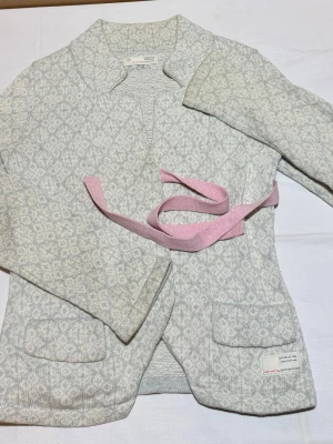 Odd Molly cardigan  - Superfin ljusgrå kofta från Odd Molly med diskret mönster och två fickor framtill. Koftan har ett rosa knytband i midjan och är tillverkad i mjukt material, perfekt för lager-på-lager. Klassisk och stilren design som passar till många outfits.