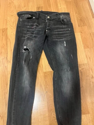 Svarta slitna jeans med hål - Säljer ett par svarta jeans med slitningar och hål på vänster lår. Jeansen har en klassisk femficksmodell och raka ben. Snyggt tvättade detaljer och en cool, avslappnad look. Perfekta för dig som gillar streetstyle och en edgy vibe.