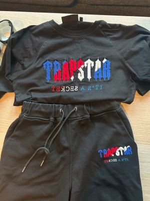 Trapstar svart set med tryck - Säljer ett svart set från Trapstar med färgglatt tryck i rött, vitt och blått på både tröjan och byxorna. Setet består av en t-shirt och shorts med dragsko i midjan. Materialet är mjukt och bekvämt, perfekt för en avslappnad stil.
