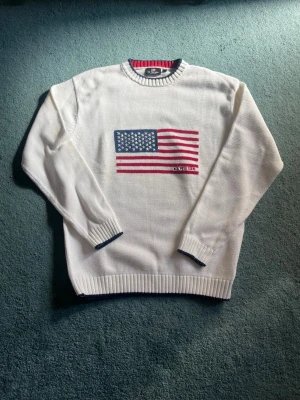 Vit stickad tröja polo assn - Vit stickad tröja från polo assn med amerikansk flagga broderad på bröstet. Tröjan har rund halsringning och mörkblå detaljer vid muddar och nederkant. Perfekt för dig som gillar klassisk och preppy stil.