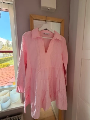 Ljusrosa långärmad klänning - Söt ljusrosa klänning med lång ärm och v-ringning. Klänningen har en avslappnad passform, krage och volangdetaljer i kjolen. Perfekt för vår och sommar. Tillverkad i mjukt bomullstyg.