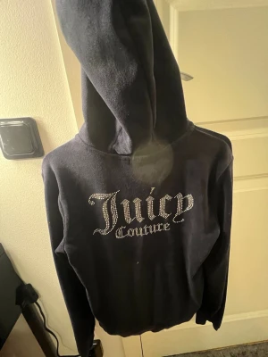 Svart Juicy Couture hoodie med strass - Cool svart hoodie från Juicy Couture med stor huva och blingade strassbokstäver på bröstet. Mjuk och skön, perfekt för att sticka ut med en ikonisk look. Materialet är fleeceliknande och ger en lyxig känsla. Snyggt broderad logga framtill.
