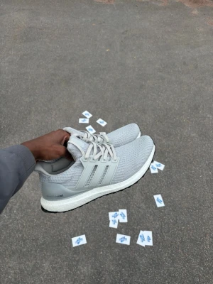 Grå Adidas Ultraboost sneakers - Säljer ett par grå Adidas Ultraboost sneakers med stickad ovandel och klassiska tre ränder på sidan. Skorna har en vit Boost-sula och svart yttersula. Perfekt för dig som gillar stilrena och bekväma sneakers.