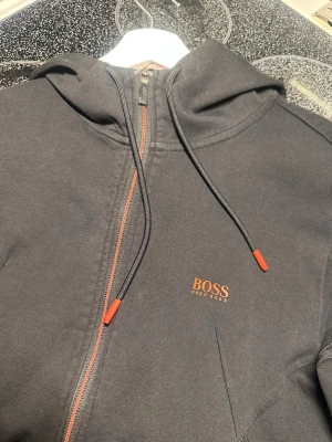 Svart hoodie från BOSS med dragkedja - Snygg svart hoodie från BOSS med dragkedja och orange detaljer på snören och logga. Klassisk huva och fickor framtill. Tillverkad i mjukt bomullsmaterial som är skönt att bära. Perfekt för en avslappnad och stilren look.