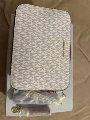 Michael Kors väska - Snygg beige handväska från Michael Kors med geometriskt mönster och guldfärgad logotyp på framsidan. Väskan har en rektangulär form och detaljer i guld. Perfekt för dig som gillar stilrena accessoarer.