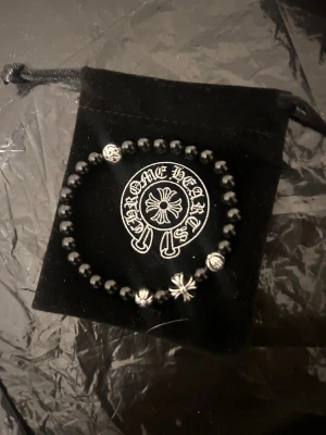 Chrome Hearts pärlarmband svart - Snyggt pärlarmband från Chrome Hearts med svarta pärlor och silverfärgade detaljer, inklusive ikoniska kors och mönstrade pärlor. Armbandet har en rund form och levereras i en svart påse med logotyp. Perfekt för dig som gillar exklusiva accessoarer.