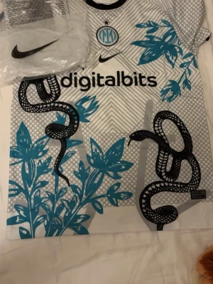 Inter Milan x Nike matchtröja ormmönster - Unik Inter Milan matchtröja från Nike med svart ormmönster och blå blommor på vit botten. Tröjan har korta ärmar, rund hals och digitalbits-logga framtill. Perfekt för fotboll eller samlare av sportkläder.