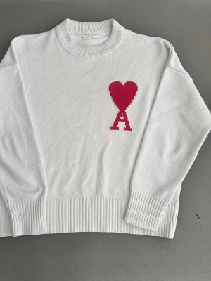 Ami Paris tröja  - Vit stickad tröja med rund hals och ribbade muddar. På bröstet finns ett rött hjärta ovanför ett stort A, vilket ger en cool och trendig look. Perfekt för dig som gillar stilrena plagg med en twist. Materialet är mjukt och bekvämt.