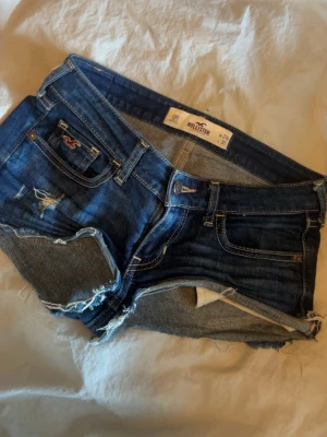 Blå jeansshorts från Hollister - Säljer ett par klassiska blå jeansshorts från Hollister med råa kanter och slitna detaljer. Shortsen har fem fickor, låg midja och en snygg tvättad look. Perfekta för varma dagar och festivaler.