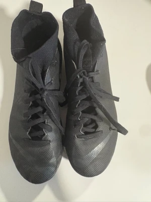 Svarta Nike fotbollsskor med dobbar - Säljer ett par svarta Nike fotbollsskor med högt skaft och snörning. Skorna har klassisk Nike-logga på sidan och dobbar på sulan för bra grepp på planen. Materialet är syntetiskt och skon har en modern, strömlinjeformad design. Säljer dessa då jag inte kör sporten längre. (De finns även knäskydd som man får gratis, men det är använda) nypris 1200kr!