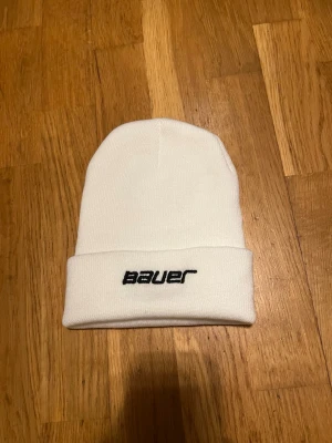 Vit Bauer mössa med broderad logga - Snygg vit mössa från Bauer med svart broderad logga framtill. Klassisk ribbstickad modell med uppvikt kant, perfekt för att hålla värmen. Passar dig som gillar sportig och clean stil.