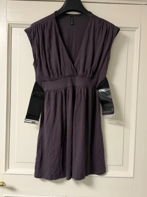 Mörklila omlottklänning med bälte - Snygg mörklila kortklänning från H&M med omlott-v-ringning och bred midja. Klänningen har ett svart bälte med silverdetalj som markerar midjan. Perfekt för dig som gillar en stilren och trendig look.