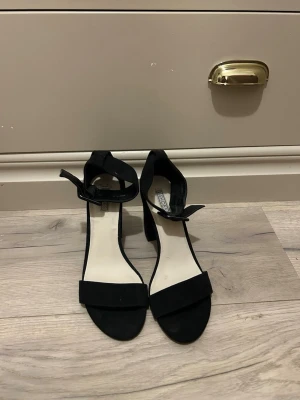 svarta klackar från nelly - Stilrena svarta pumps från Nelly med öppen tå, bred klack och ankelrem med rosett. Skorna är i mockaimitation och har en klassisk, elegant design som passar perfekt till många olika outfits.