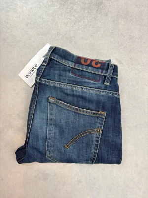 Blå jeans från Dondup med raka ben - Snygga blå jeans från Dondup med klassisk femficksmodell och raka ben. Jeansen har slitningar och tvättade detaljer för en cool look. Gula kontrastsömmar och patch med röd detalj bak i midjan. Perfekta för dig som gillar en avslappnad men trendig stil.