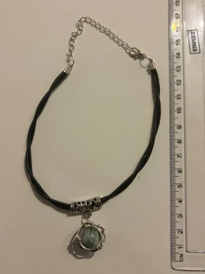 Choker med grön pärla och svart band - Unikt halsband med svart tvinnat band och en grön pärla infattad i ett dekorativt silvrigt hänge. Halsbandet har justerbar kedja och detaljer i metall med mönster. Perfekt för dig som gillar boho eller alternativ stil.