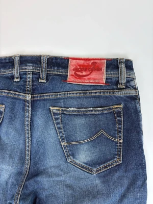 Blå jeans från Jacob Cohen - Snygga blå jeans från Jacob Cohen med klassisk femficksdesign, kontrastsömmar och en röd patch bak i midjan. Jeansen har en lätt tvättad look och diskreta slitningar på låret. Tillverkade i denim med rak passform.