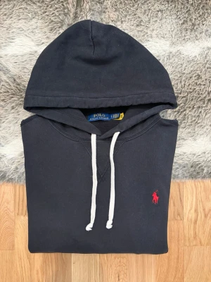 Svart Polo Ralph Lauren Hoodie - Storlek L - Hej! 👋 Säljer en riktigt snygg och klassisk svart Polo Ralph Lauren hoodie i storlek L. Den har den ikoniska röda pony-loggan broderad på bröstet 🐎 – en tidlös färgkombination som alltid går hem! Tröjan är i mycket gott skick, välvårdad och känns fräsch. ✨ 100% äkta med både tvättlappar och QR-kod kvar för enkel verifiering ✅. Ett perfekt premium-basplagg som håller stilen år efter år. Fraktar snabbt! 🚛📦💬💯 