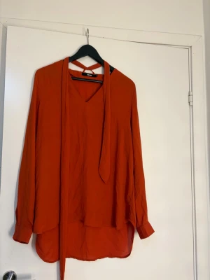 Rostfärgad blus med knytband - Trendig rostfärgad blus från H&M med v-ringning och långa ärmar. Blusen har ett dekorativt knytband vid halsen och rundad nederkant. Tillverkad i ett lätt, flowy material som ger en avslappnad men stilren look.