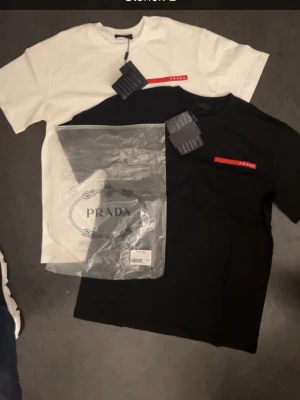 Prada t-shirt svart och vit - Två stilrena t-shirts från Prada, en i svart och en i vitt. Båda har korta ärmar och en röd logodetalj på bröstet. Klassisk passform och tillverkade i mjuk bomull. Perfekta för en clean och trendig look. En för 400kr båda för 800kr