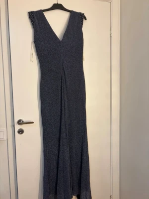 Blå glittrig långklänning med slits - Snygg blå långklänning med glittrigt tyg och djup v-ringning både fram och bak. Klänningen har en slits framtill och dekorativa pärlor vid axlar och midja. Perfekt för speciella tillfällen när du vill sticka ut.