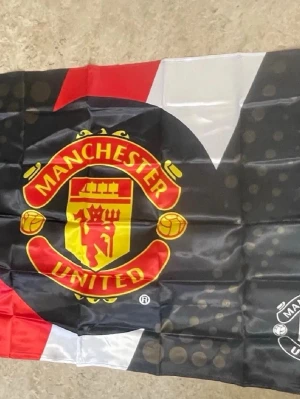 Manchester United supporterflagga - Stor supporterflagga med Manchester Uniteds klassiska klubbmärke i rött, gult och svart. Flaggan har även vita och röda detaljer och är tillverkad i ett lätt tygmaterial. Perfekt för att visa sitt stöd för laget.
