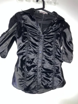 Svart glansig blus med rynk - Snygg svart blus i glansigt material med markerade rynkdetaljer framtill och baktill. Blusen har krage, knäppning framtill och trekvartsärm. Perfekt för dig som vill ha en lite extra statement-blus till din outfit.