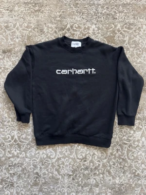 Svart sweatshirt från Carhartt - Säljer en svart sweatshirt från Carhartt med broderad vit logga på bröstet. Tröjan har rund halsringning, ribbade muddar vid ärmslut och nederkant samt en avslappnad passform. Perfekt för en casual och streetwear-inspirerad stil.