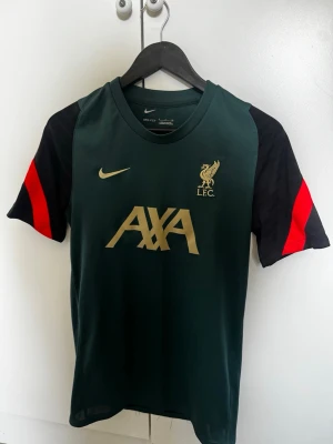 Liverpool tränings t-shirt Nike - Mörkgrön Liverpool FC tränings t-shirt från Nike med svarta ärmar och röda detaljer. Framsidan har guldiga loggor för Nike, LFC och AXA. Tillverkad i Dri-Fit material som andas, perfekt för träning och sport.