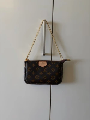 handväska - Snygg liten handväska  i klassiskt monogrammönster. Väskan är brun med guldiga detaljer och kedja som axelrem. Tillverkad i canvas med läderdetaljer och dragkedja upptill. Perfekt för att bära det viktigaste med stil.