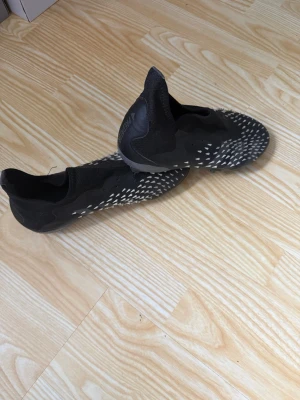 Adidas Predator svart fotbollssko - Säljer ett par svarta Adidas Predator fotbollsskor med vita detaljer och mönstrad ovansida. Skorna har strumpliknande skaft och fasta dobbar för spel på gräs. Snygg och modern design som ger bra grepp och kontroll på planen.