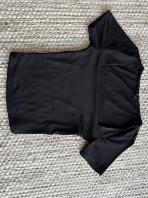 Svart kortärmad topp med fyrkantig ringning - En stilren svart topp med korta ärmar och en modern fyrkantig ringning. Tillverkad i ett glansigt och stretchigt material som sitter snyggt på kroppen. 