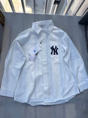 Vit skjorta New Era - Säljer en vit skjorta från MLB med klassisk krage och långa ärmar. Skjortan har subtila ränder i tyget och en stor, mörkblå NY Yankees-logga broderad på bröstfickan. Perfekt för dig som gillar sportig stil och vill sticka ut.
