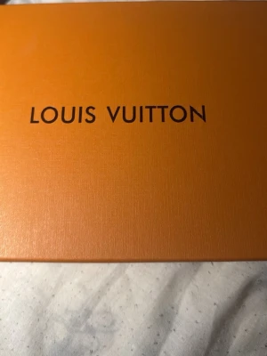 Louis Vuitton orange box - Exklusiv orange box från Louis Vuitton med svart logga på locket. Boxen har en stilren och lyxig design, perfekt för att förvara accessoarer eller använda som presentask. Materialet är hård kartong med lätt struktur.