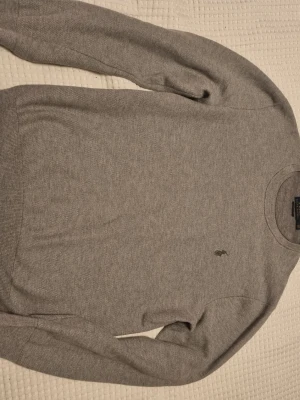 Grå stickad tröja från Polo Ralph Lauren - Snygg grå stickad tröja från Polo Ralph Lauren med klassisk rund halsringning och diskret broderad logga på bröstet. Tröjan har långa ärmar och ribbade muddar vid ärmslut och nederkant. Perfekt för lager-på-lager och stilrena outfits.