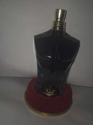 Jean Paul Gaultier Le Male parfym - Unik parfymflaska från Jean Paul Gaultier formad som en manlig torso i mörkblått glas med guldfärgad kork. Flaskan står på en rund röd sammetsliknande bas med gulddetaljer. En ikonisk doft som sticker ut i badrumshyllan. Cirka halva kvar.