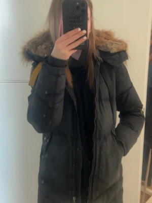 Svart lång parajumper jacka med fuskpäls - Svart lång parajumper jacka med luva och mycket päls på luvan. Jackan har en dragkedja framtill som gått av men finns i jackfickan så man kan fixa det , fickor på sidorna och en patch på ärmen. Jackan kan behöva kemtvättas den ser mer vit ut på sista bilden en vad den är i verkligheten. Perfekt för kalla dagar med sin varma och mysiga känsla. 