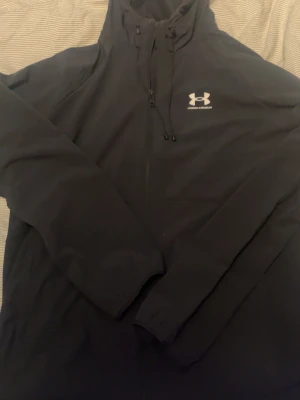 Svart träningsset från Under Armour - Snyggt svart träningsset från Under Armour med jacka och byxor. Jackan har dragkedja, huva med dragsko och logga på bröstet. Byxorna har resår i midjan och logga på låret. Perfekt för löpning eller gym. Materialet är lätt och smidigt.