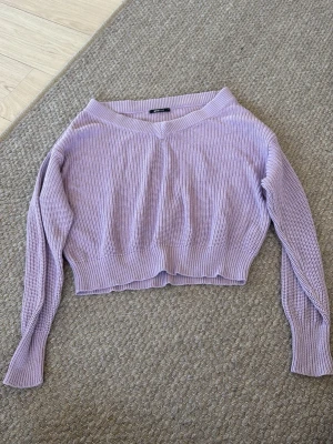 Ljuslila stickad cropped tröja - Säljer en ljuslila stickad tröja med cropped passform och bred ribbad kant. Tröjan har långa ärmar och en rund halsringning. Perfekt för att skapa en avslappnad och trendig look.