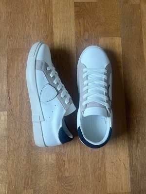 Vita sneakers från Philippe Model - Snygga vita sneakers från Philippe Model med grå detaljer och mörkblå häl. Skorna har klassisk låg profil, vita skosnören och en robust gummisula. Materialet är en mix av läder och mocka, vilket ger en stilren och modern look.
