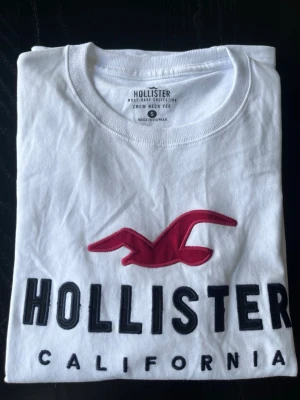 Vit Hollister tröja - Snygg vit tröja från Hollister med stor röd fågellogga och svart text 'HOLLISTER CALIFORNIA' på bröstet. Klassisk rund halsringning och är lång armad. Tillverkad i mjuk bomull för en bekväm känsla.