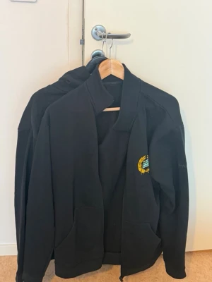 Hammarby tröja - Svart hoodie med dragkedja och Hammarby IF:s klassiska klubbmärke broderat på bröstet. Tröjan har två fickor framtill och en stilren design som passar perfekt för dig som vill visa ditt stöd för laget. Materialet är mjukt och bekvämt.