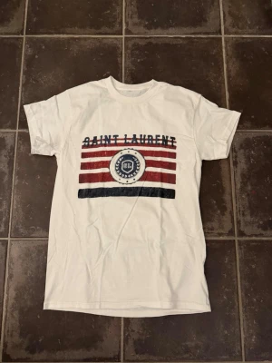 Saint Laurent t shirt  - Riktigt fet och eftertraktad t-shirt med Saint Laurent loggan i storlek S. Väldigt bra skick!  Obs: Jag är lite osäker på äktheten då lappen i nacken saknas, därför säljs den billigare (499 kr, nypris ca 3000 kr).  längd cirka 65cm, bredd cirka 44cm. Tveka inte att höra av dig vid frågor eller funderingar!