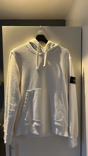 Vit hoodie från Stone Island - Säljer en clean vit hoodie från Stone Island med klassisk huva och dragsnören. Hoodien har en stor magficka och den ikoniska svarta logotypen på vänster ärm. Tillverkad i mjukt bomullsmaterial som är skönt mot huden. Perfekt för dig som gillar streetwear och enkel stil.