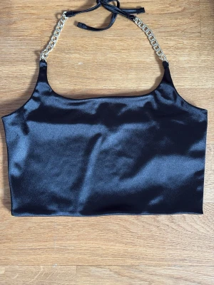 Svart satin bandeau topp med kedjor - Snygg svart bandeau topp från FB Sister i glansigt satinmaterial. Toppen har guldfärgade kedjor som axelband och knyt i nacken för extra detalj. Perfekt för dig som vill sticka ut med en trendig och unik topp.