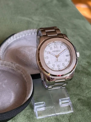 Seiko automatisk klocka i stål - Stilren Seiko klocka med automatisk urverk och vit urtavla. Klockan har tydliga siffror, datumvisning och klassisk design som passar till många stilar. Perfekt för dig som gillar tidlösa accessoarer.