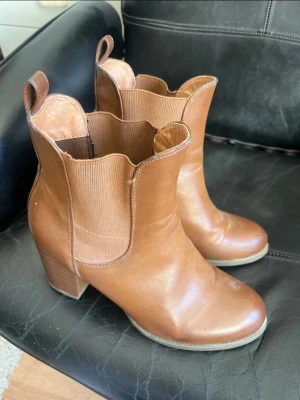 Bruna Chelsea boots med klack - Snygga bruna Chelsea boots i läderimitation med bred resår på sidorna och markerad klack. De har rundad tå och dragflikar upptill för enkel påtagning. Perfekta för dig som vill ha en klassisk men trendig look.